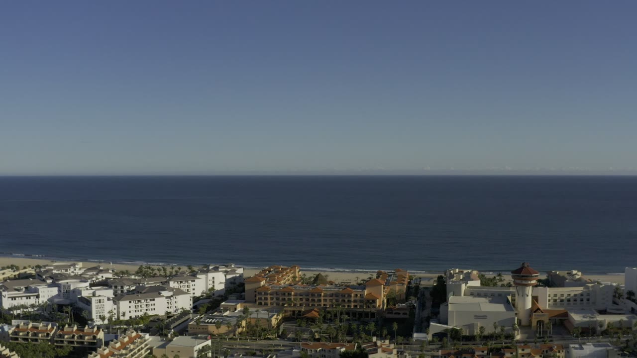 Opening aerial frame over Los Cabos, Mexico, showing clustered white and terracotta buildings, a golf course, beach, and blue ocean horizon with no visible people.