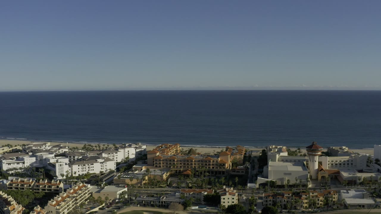 Los Cabos aerial view one second into the clip, with the ocean across the top of frame, a beach strip below it, roads and rooftops in front, and no visible people.