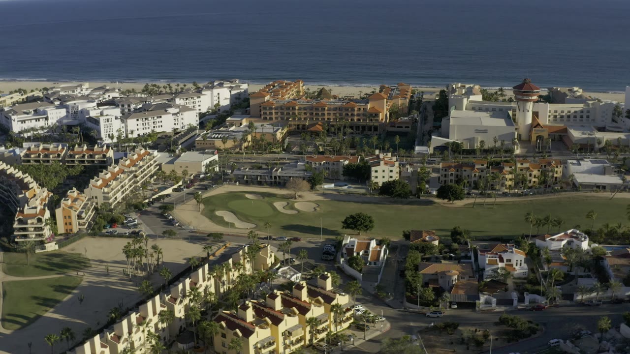 Opening aerial frame in Los Cabos, Mexico, showing ocean beyond a beach, white and tan buildings, red-tile roofs, roads, palm trees, and a golf course with no visible people.