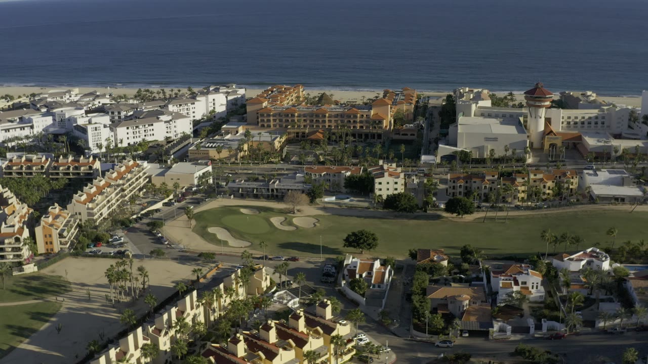 Mid-clip aerial view in Los Cabos, Mexico, showing a broad fairway, curved sand bunkers, beachside buildings, ocean water, foreground rooftops, and no visible people.