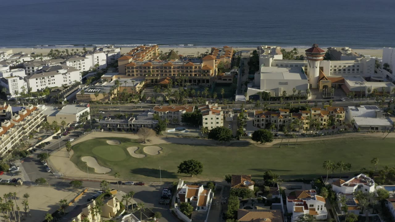 Los Cabos aerial frame showing the shoreline, white and terracotta-roofed buildings, a tower-like structure, road grid, green grounds, and no visible people.
