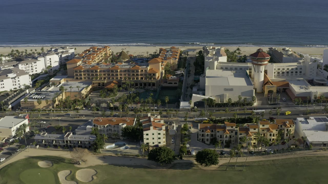 Aerial frame in Los Cabos centered on red-roofed coastal buildings and a white tower-like structure, with the beach and ocean in back and no visible people.