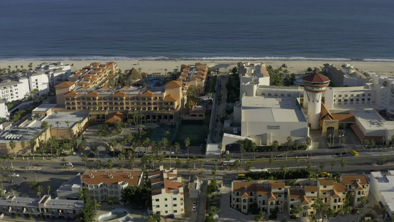 Opening aerial frame over Los Cabos, Mexico, with a wide road and small vehicles below white and terracotta buildings, sandy beach, blue ocean, and no visible people.