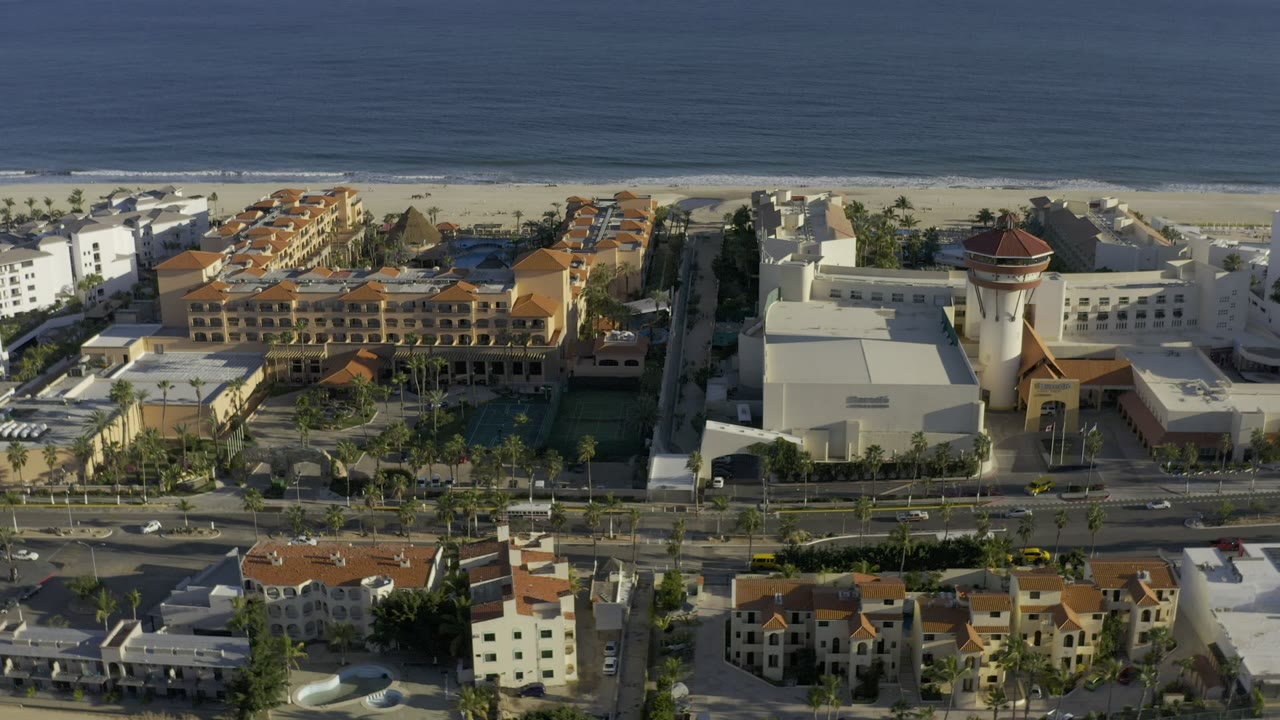 The second aerial frame keeps Los Cabos road traffic in the foreground, hotel-style buildings in the center, the beach behind them, and no visible people.
