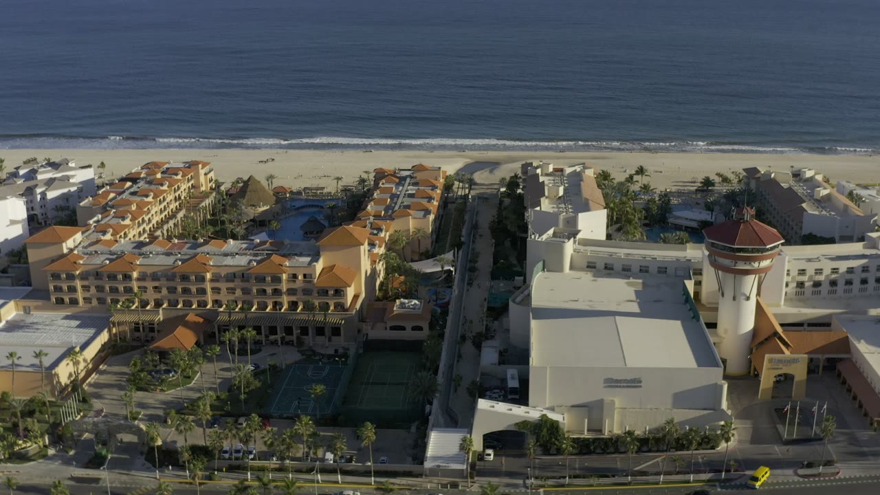 Aerial frame in Los Cabos, Mexico, with a white building and red-roofed tower in the foreground, tan buildings to the side, beach and ocean behind, and no visible people.
