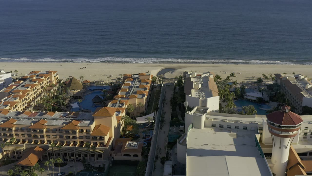 A high aerial frame over Los Cabos, Mexico shows tan resort buildings, a sandy beach, and dark blue ocean water with no visible people.
