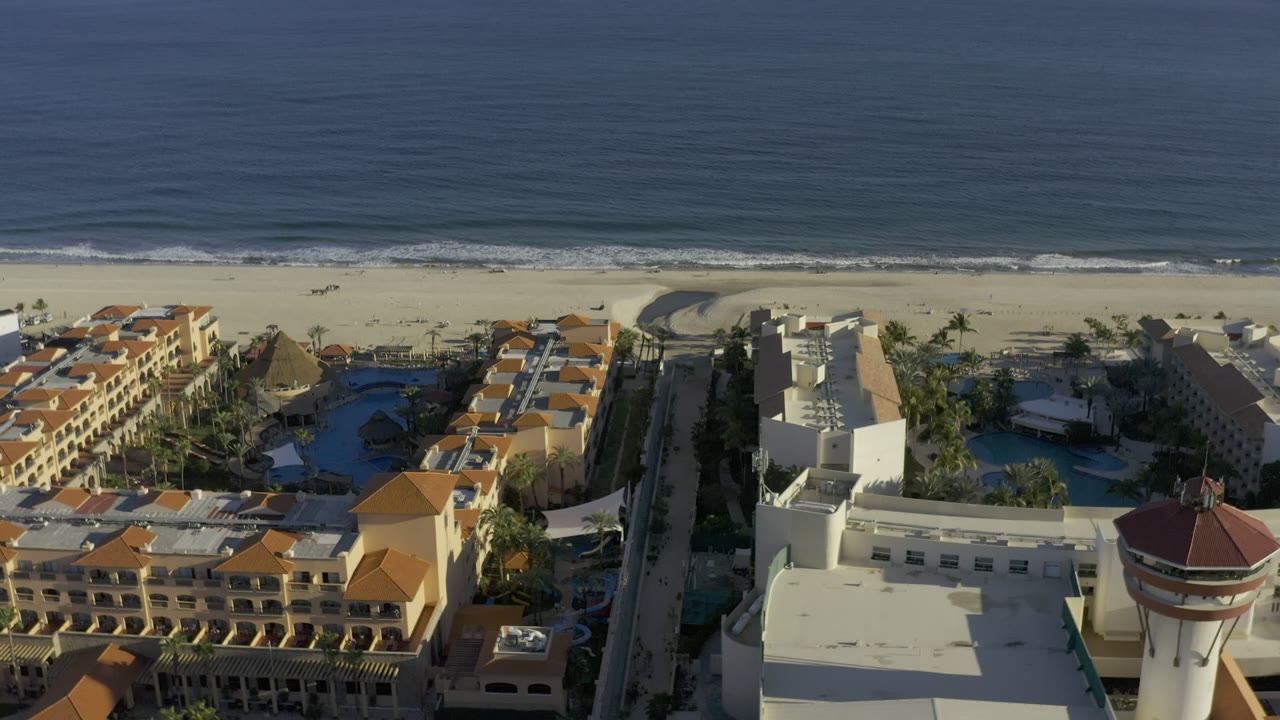 An elevated Los Cabos, Mexico view centers white and tan beachfront buildings below a band of sand and calm ocean, with no visible people.