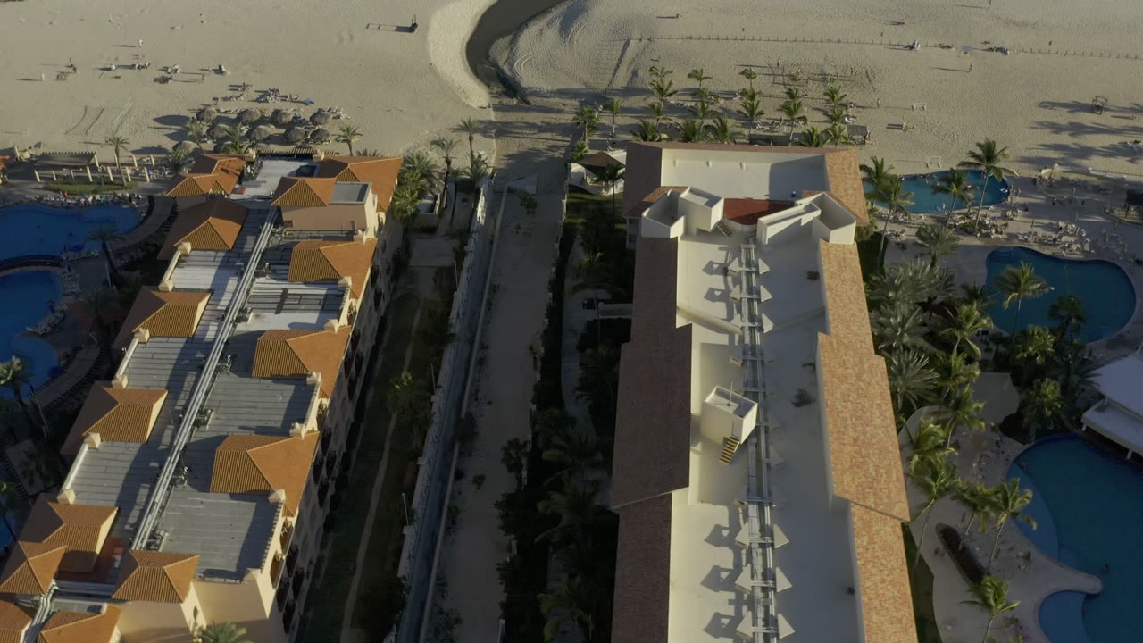 Aerial frame over Los Cabos, Mexico, with a tall cream-colored building centered between condo roofs, curved blue pools, palm trees, beach umbrellas, pale sand, and small people visible in the distance.