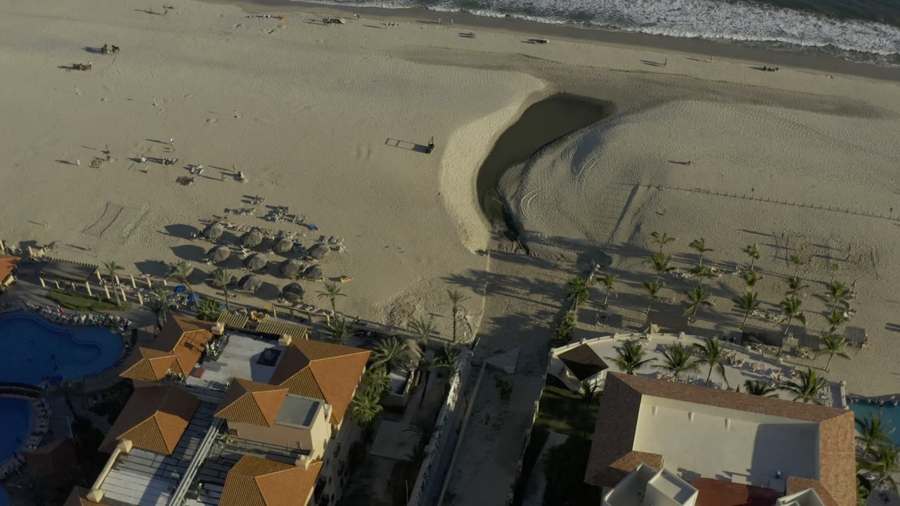 Aerial view of beach houses, sand, palms, and ocean surf.
