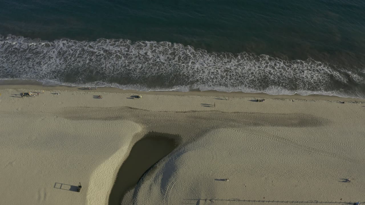 Overhead view of dark teal water and white surf meeting pale sand in Los Cabos, Mexico, with small beach objects visible and no visible people.