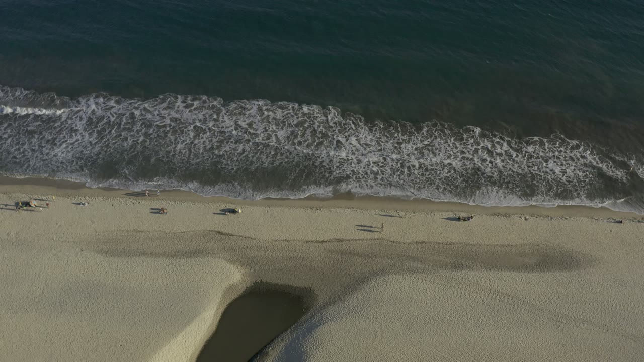 Aerial view of white water spread farther across the foreshore in Los Cabos, Mexico, with pale beach sand below and no visible people.