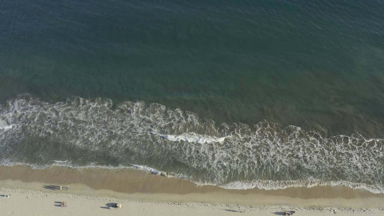 Overhead view in Los Cabos, Mexico, with teal surf and white foam meeting pale beach sand near dark tide-pool shapes, with no recognizable people visible.