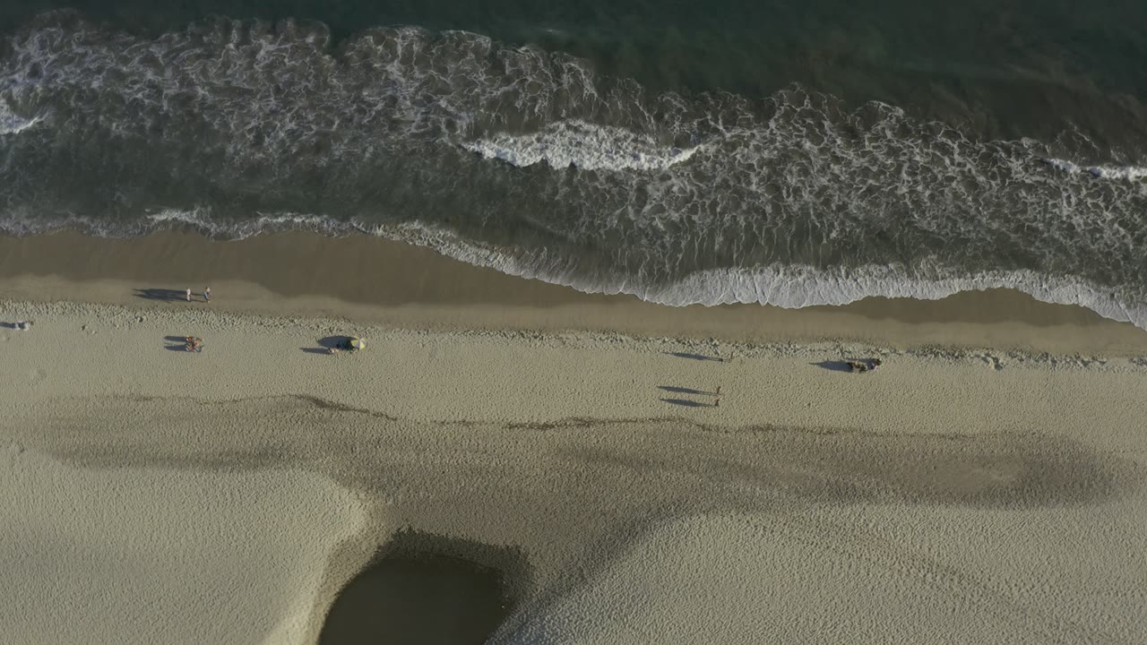 Aerial Los Cabos beach frame showing the surf line across the top, textured sand through the middle, and a dark water channel cutting into the lower beach, with no recognizable people visible.