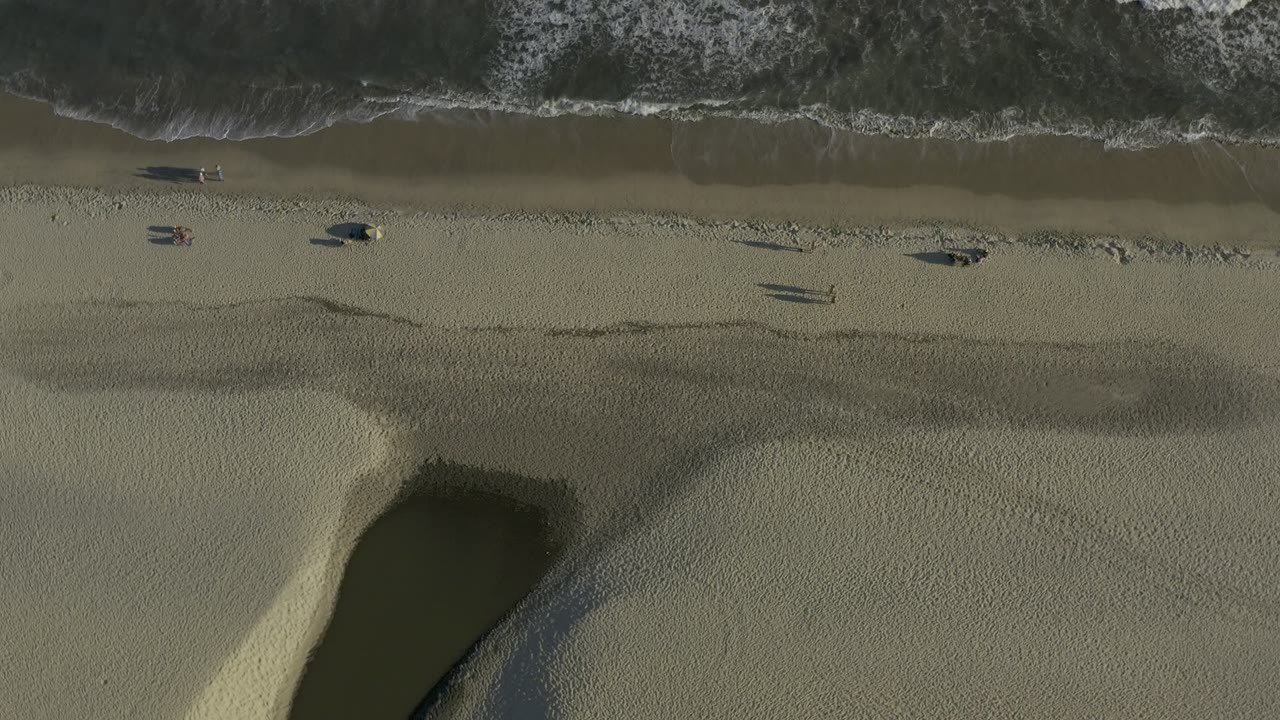 Overhead beach view with surf, pale sand, and dark tide pools.