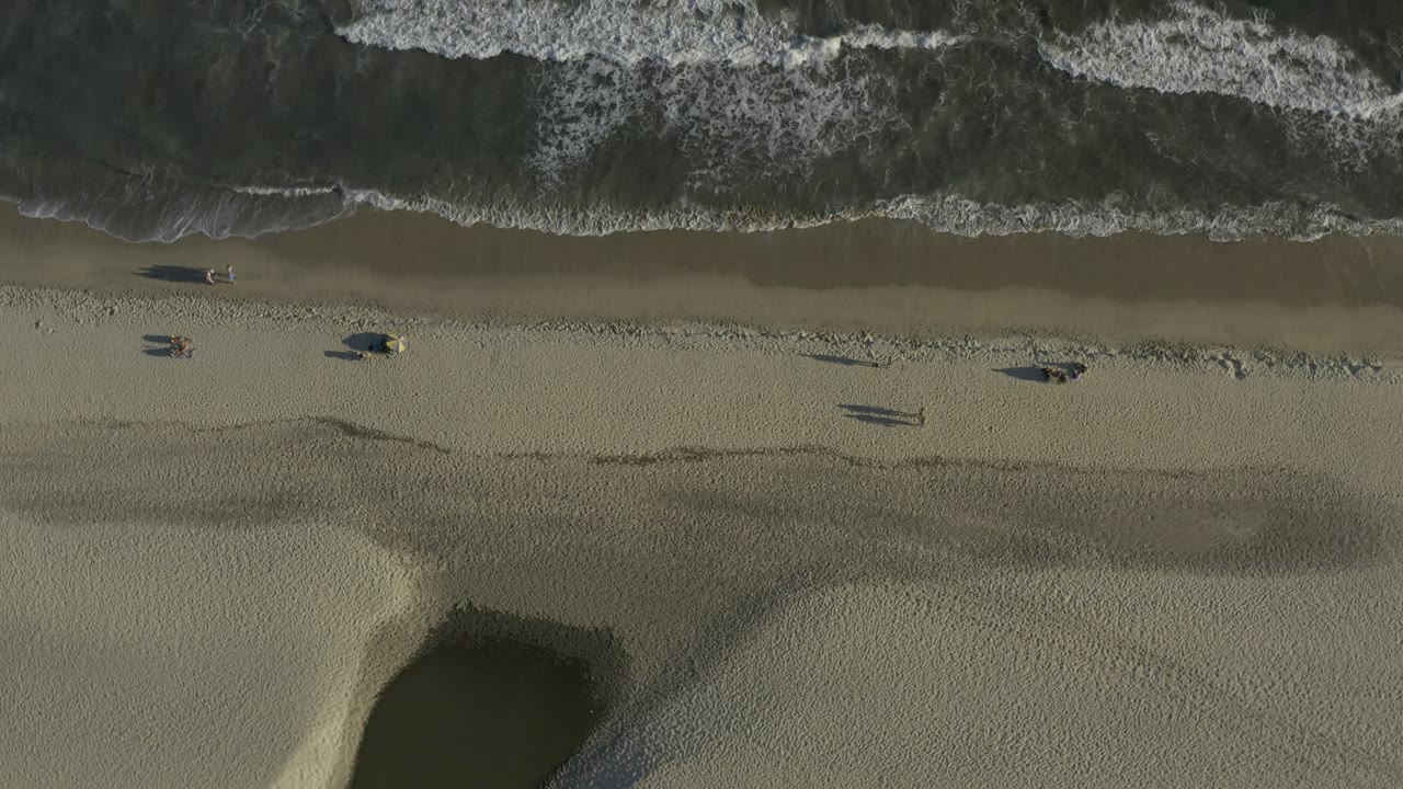 Second overhead Los Cabos frame showing foamy waves pushing toward the beach, a long wet-sand strip, small scattered figures, and dark pools cut into the lower sand.