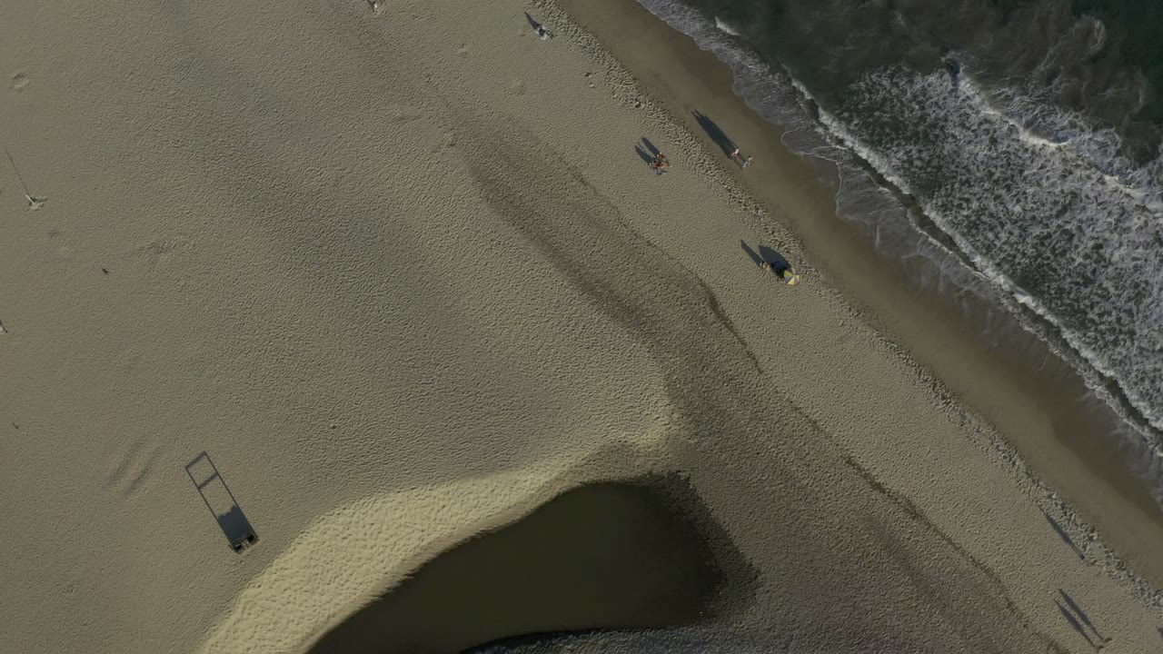Overhead view in Los Cabos, Mexico showing diagonal surf, wide sand, several small beachgoers, long shadows, and a dark water pocket at the lower edge.