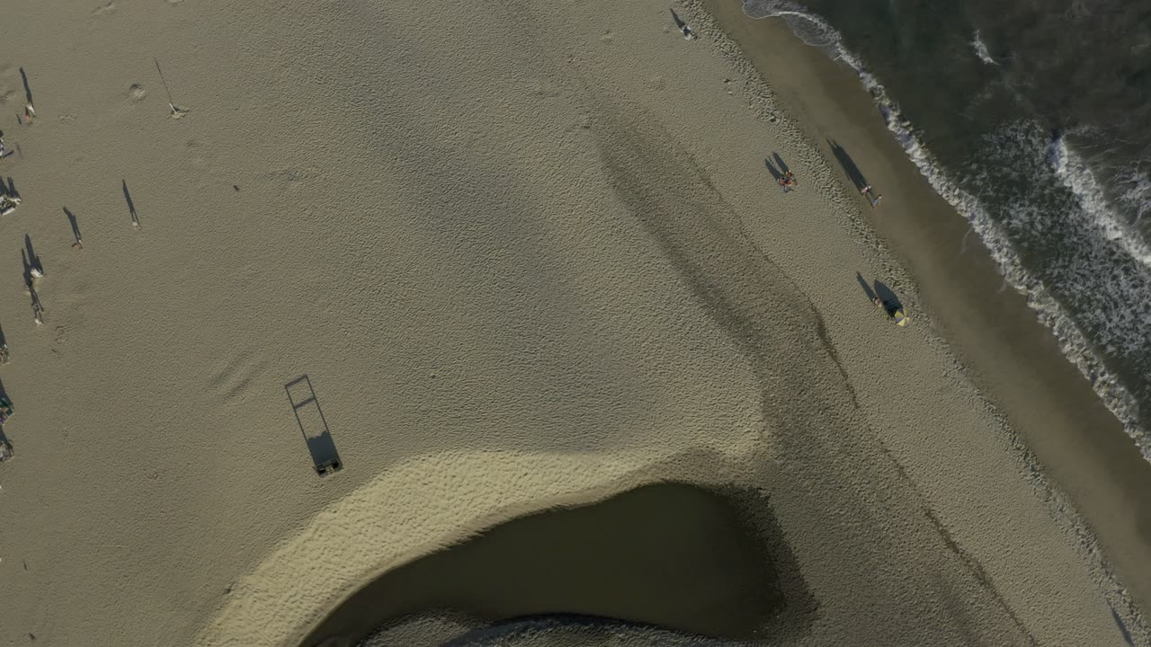 Aerial view in Los Cabos, Mexico with the surf shifted toward the top right, scattered beachgoers near the waterline, open sand, and a dark shoreline pocket below.