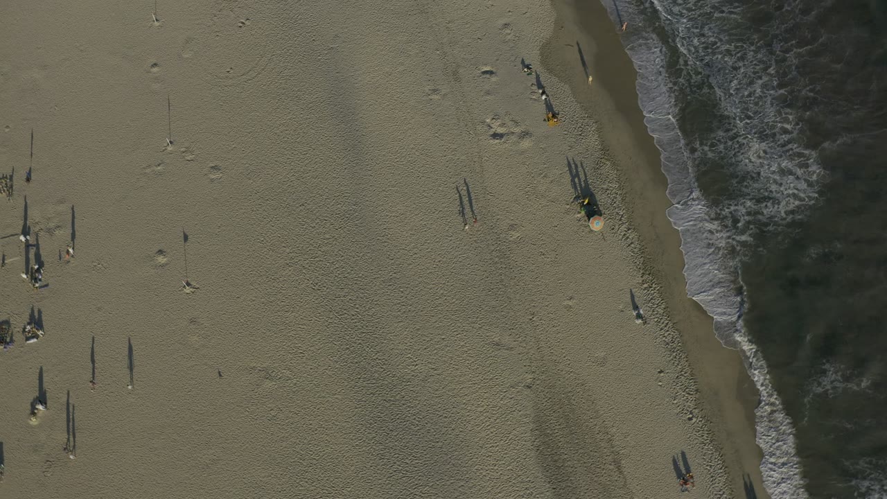 Aerial Los Cabos shoreline frame with the wave line running vertically through the image, open sand on both sides, and small distant figures that are not identifiable.