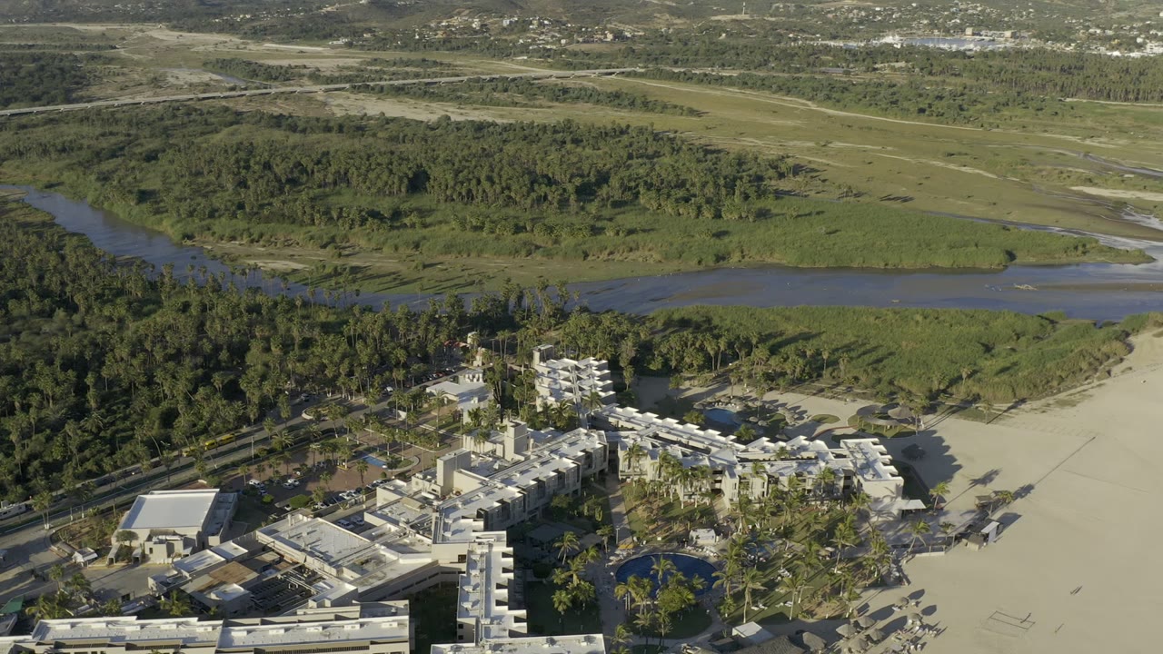 Los Cabos aerial frame with white low-rise buildings in the lower foreground, palms and sand nearby, a winding water channel beyond, and no visible people.