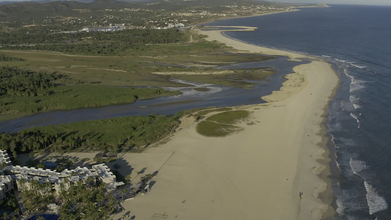 At 0:01, the Los Cabos coastline fills the frame with a pale sandbar, river water bending behind the beach, ocean surf on the right, distant hills, and no visible people.