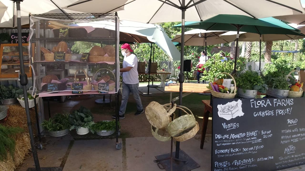 Outdoor food and vegetable stall in San Jose del Cabo with umbrellas overhead, baskets and leafy greens in front, a chalkboard produce sign, and a partially visible worker near the display.