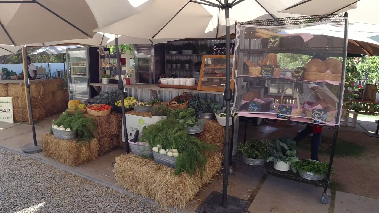 Opening view of a San Jose del Cabo outdoor food stall with hay bales, leafy greens, fruit, white umbrellas, display cases, chalkboard signs, and no visible people.