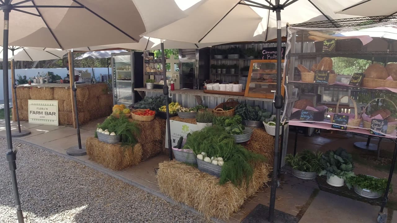 A closer angle on the San Jose del Cabo market stall shows stacked produce, hay bales, a service counter, a refrigerated display case, chalkboard menus, and no visible people.