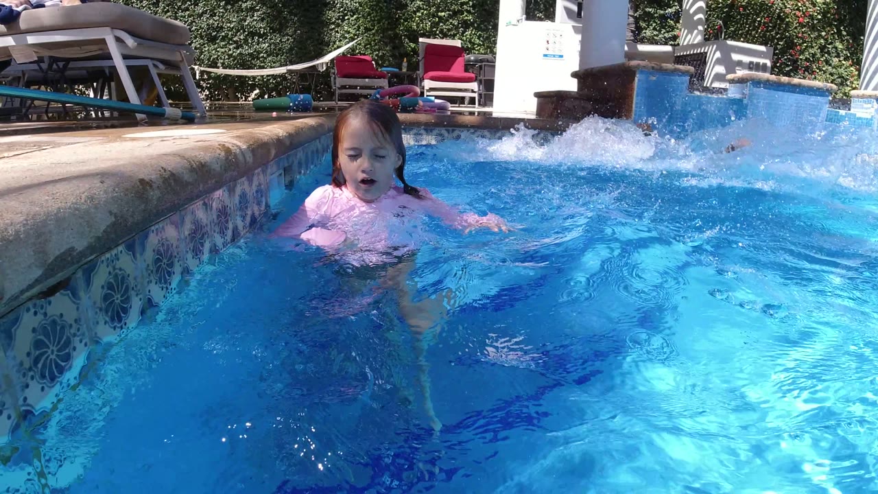 A young child in a pink swim shirt stands near the tiled edge of a sunlit swimming pool in San Jose del Cabo, Mexico, with lounge chairs and garden plants behind the water.