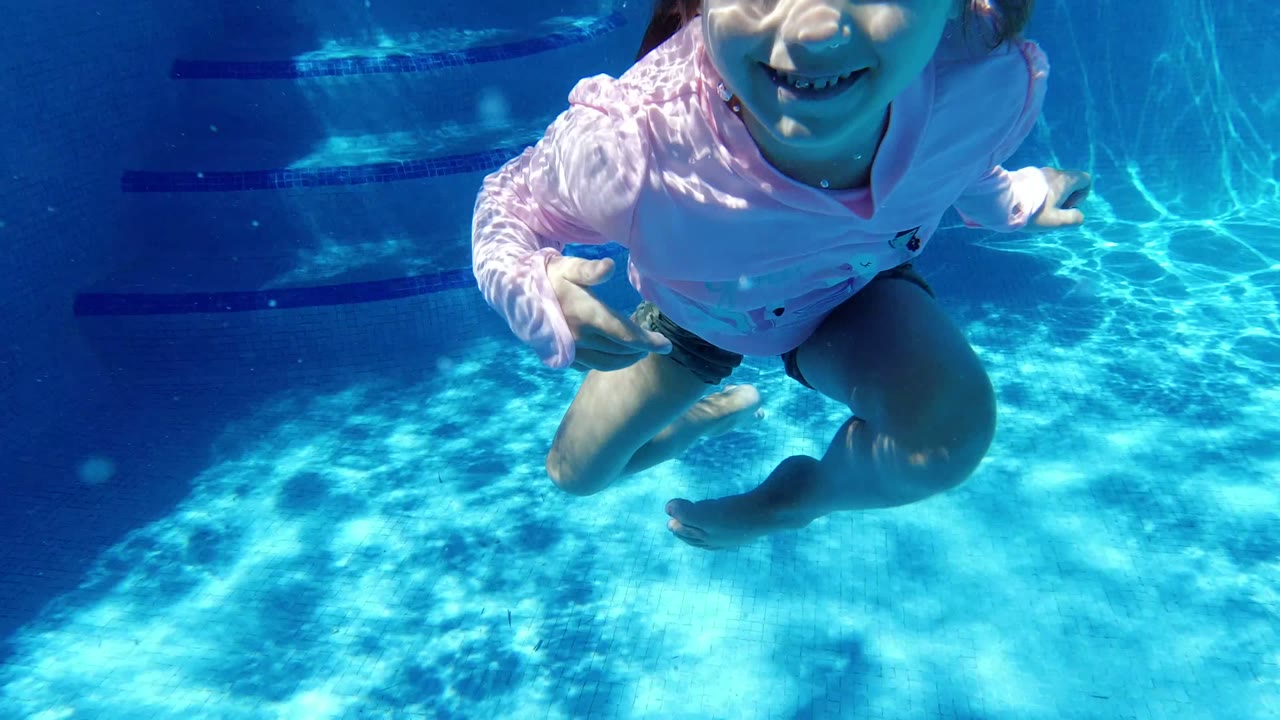 Young girl underwater in a blue pool, partly cropped near the camera.