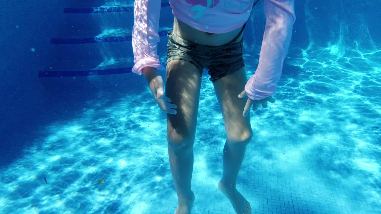The child is cropped at the head while floating upright underwater over the bright pool floor in San Jose del Cabo, Mexico.