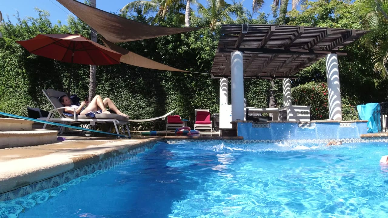 At 0:01 in San Jose del Cabo, the poolside composition shows an adult reclined under sun, a dog near the waterline, red seating, shade sails, and dense greenery behind the pool.