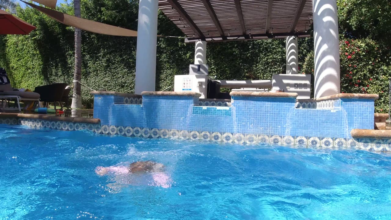 A child is mostly underwater in a blue swimming pool in San Jose del Cabo, Mexico, with lounge chairs, white columns, and greenery behind the pool edge.