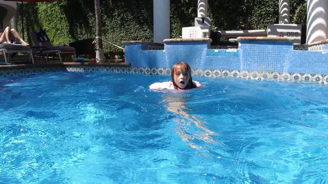 A child faces the camera while treading in the bright pool water in San Jose del Cabo, Mexico, with a lounge area and a background person on a chair.