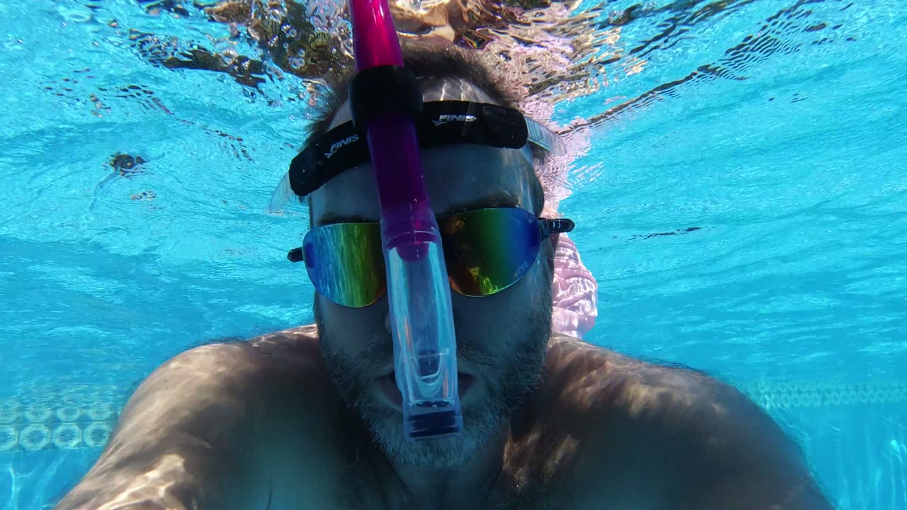 A man in San Jose del Cabo, Mexico remains centered underwater, with stronger surface ripples above his head and turquoise pool water filling the frame.