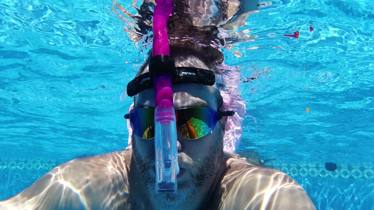 Man in snorkel gear close to the camera underwater in a bright blue pool.