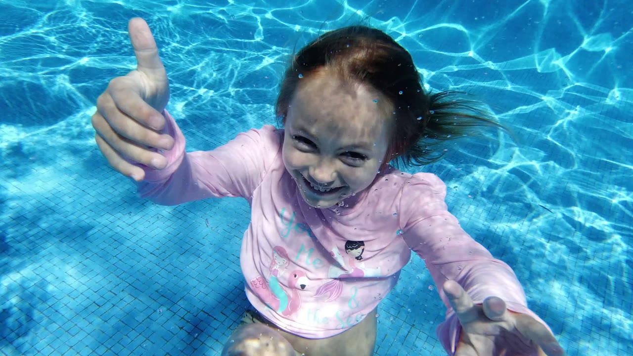 Young girl underwater in a clear blue pool in San Jose Del Cabo, Mexico, smiling near the camera with one hand giving a thumbs-up.