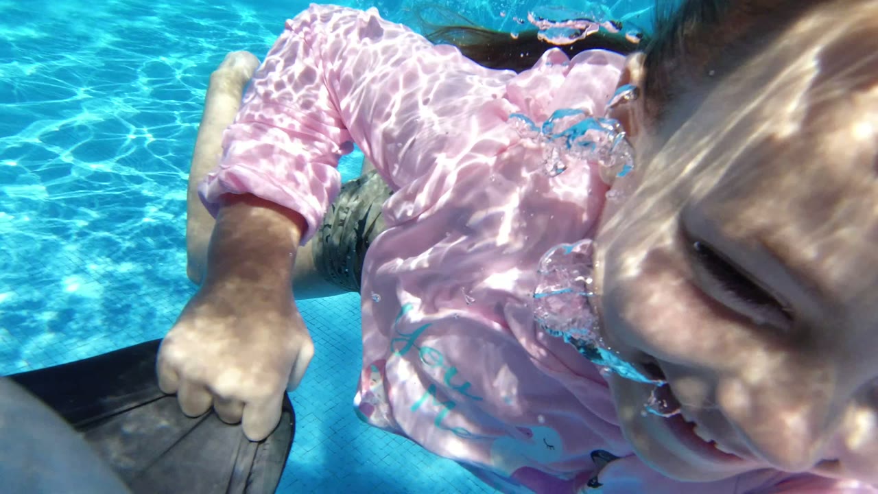 Child in pink swim shirt underwater beside an adult arm in a blue pool.