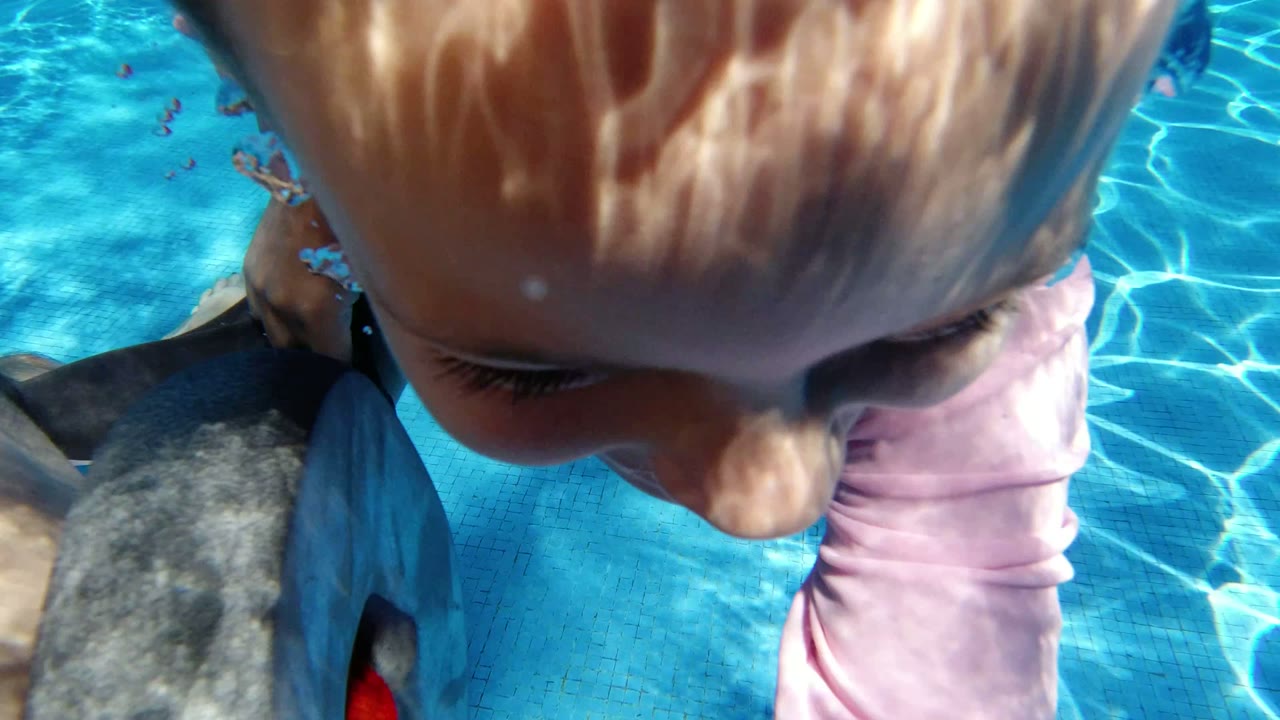 A close pool-surface view shows the child's head and pink sleeve near an adult arm in clear blue water in San Jose Del Cabo, Mexico.