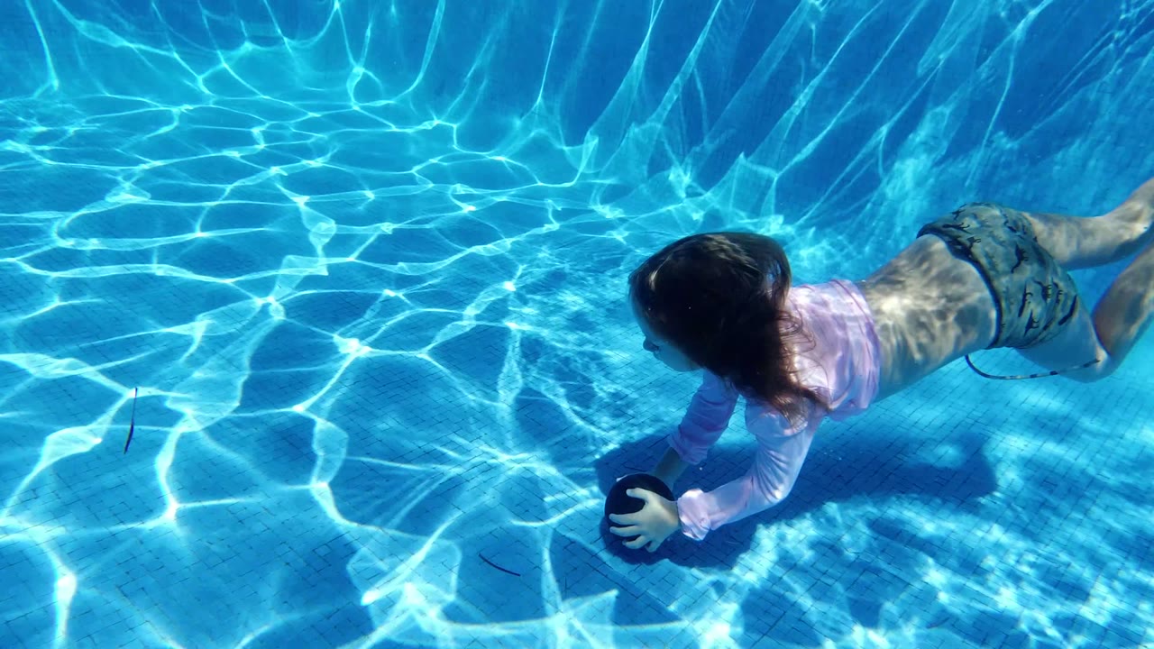 Girl in pink swim shirt reaching for a dark ball in a blue pool.