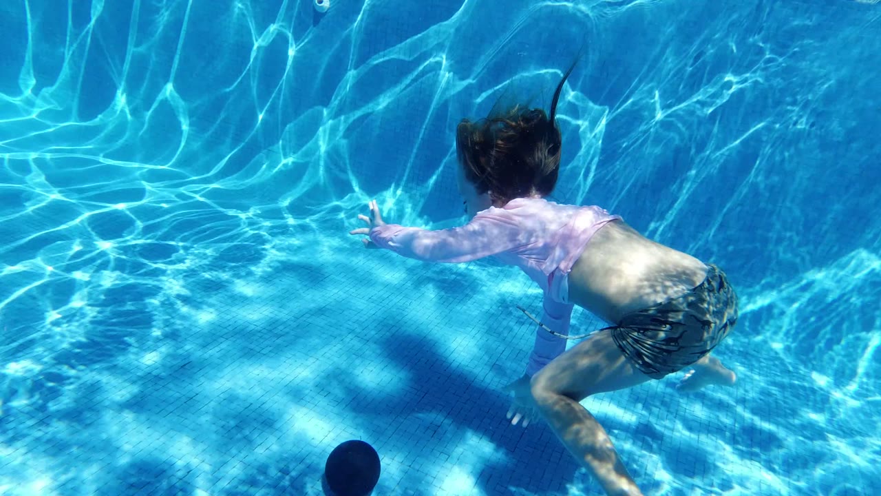 Child underwater in a blue pool reaching toward a dark round object.