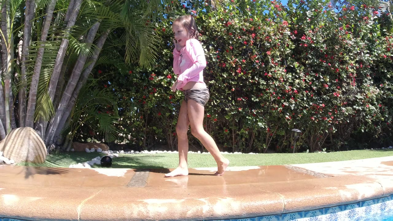 Child walking barefoot beside a pool with tropical plants behind.