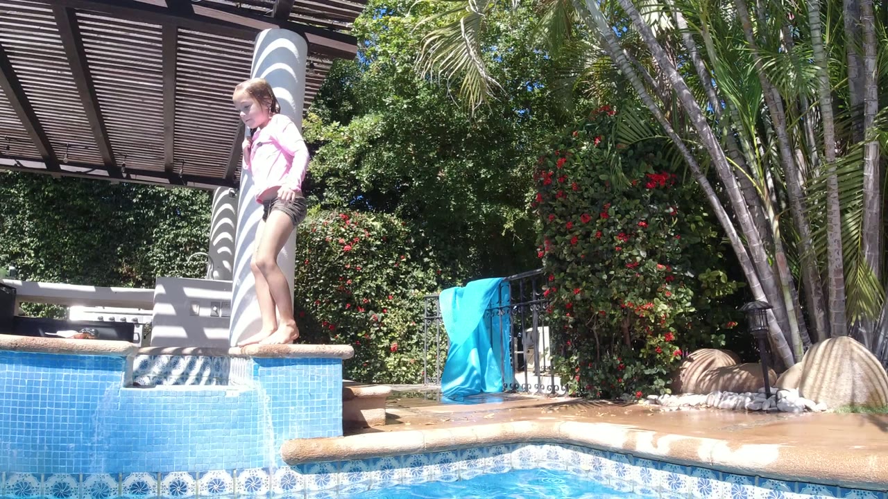 A young girl stands upright on the raised blue pool edge in San Jose del Cabo, Mexico, with striped shade from a pergola and palms in the background.
