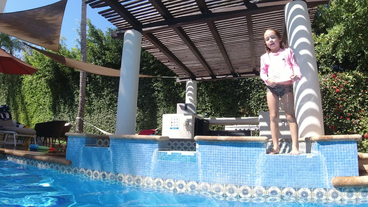 Young girl in a pink top stands on the blue tiled pool edge in San Jose del Cabo, Mexico, with sunlit water in front and shaded seating behind her.