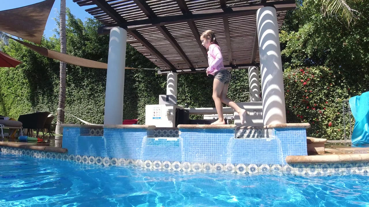 Girl in pink walks to the right along the poolside ledge in San Jose del Cabo, Mexico, with a white column and shaded patio furniture behind her.