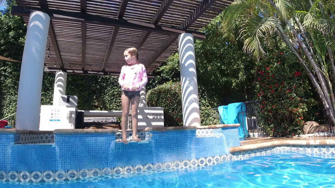 In San Jose del Cabo, Mexico, the child stands upright on the pool ledge and looks to the side, with sunlight reflecting off the water and white pergola columns behind.
