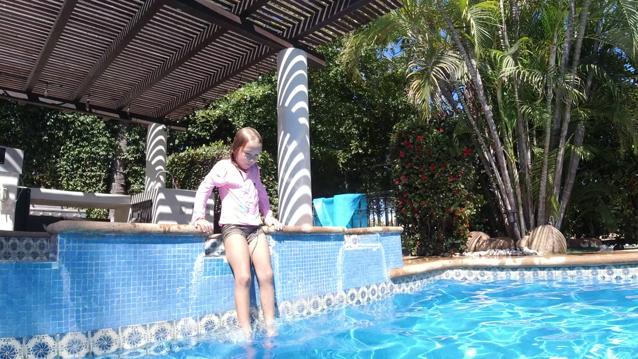A child in a pink top sits on the blue-tiled edge of a resort pool in San Jose del Cabo, Mexico, with feet in the water and sunlit ripples in the foreground.