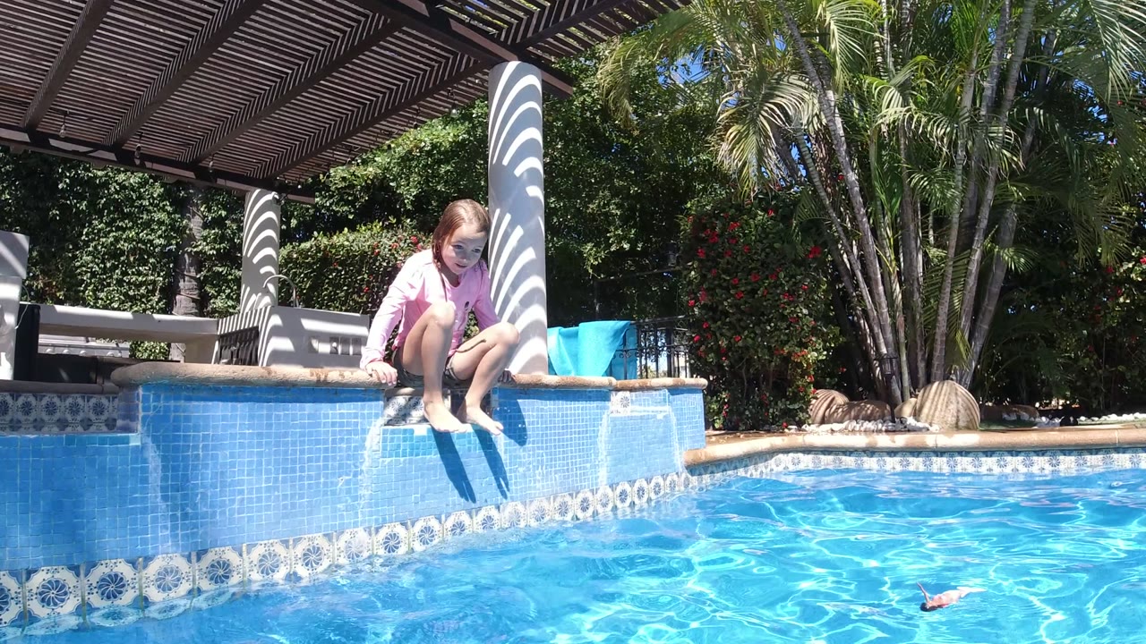 The child crouches forward on the raised blue pool wall in San Jose del Cabo, Mexico, with bare feet near the water and a blue bucket visible on the right.