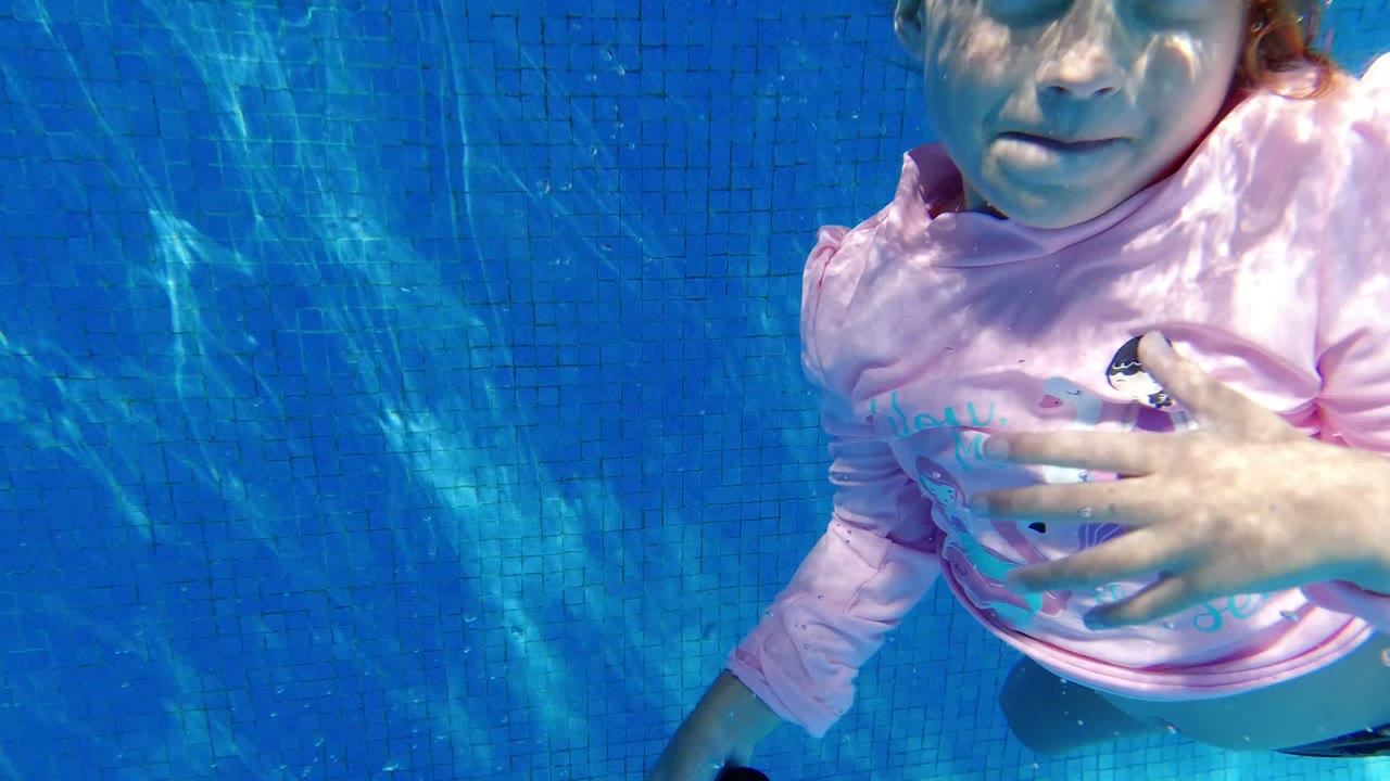 In San Jose del Cabo, Mexico, the girl faces the camera underwater with one hand near her chest, blue pool tiles behind her, and sunlight ripples across her face and shirt.