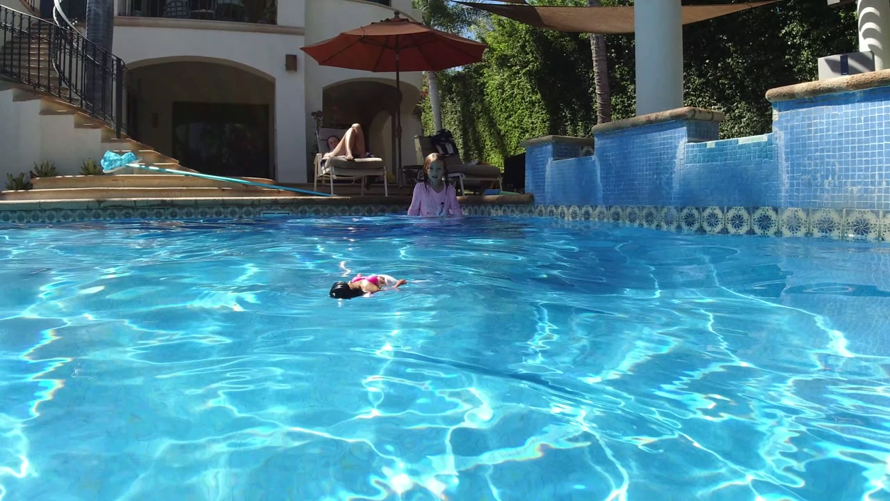Child floating in a blue swimming pool with adults near the patio behind.