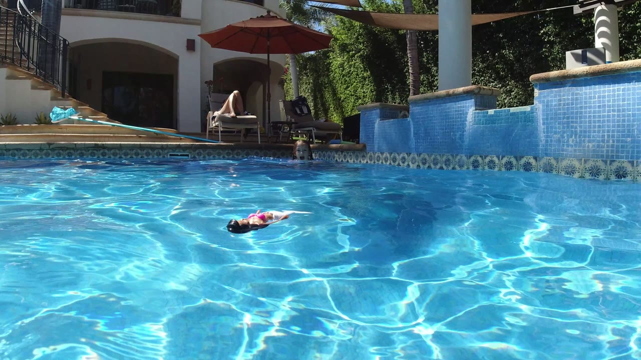 At 0:01 in San Jose del Cabo, rippled pool light surrounds the child and doll as a seated adult remains under a red umbrella near the house patio.